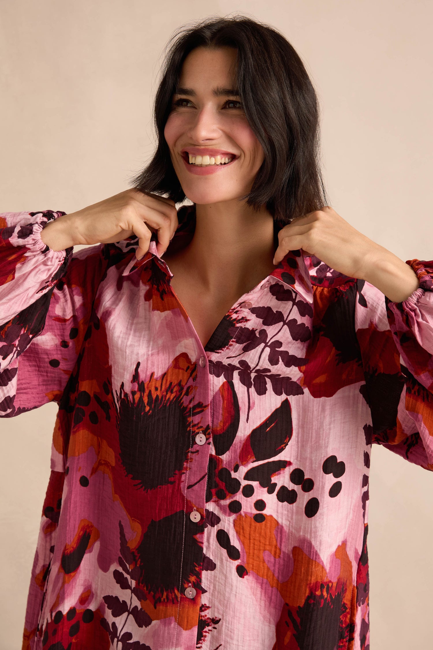 A woman smiles as she adjusts the collar of the Crinkle Viscose Shirt, a longline, button-up style featuring a bold pink, red, and black botanical print and crafted from soft crinkle viscose. She stands indoors against a neutral background.