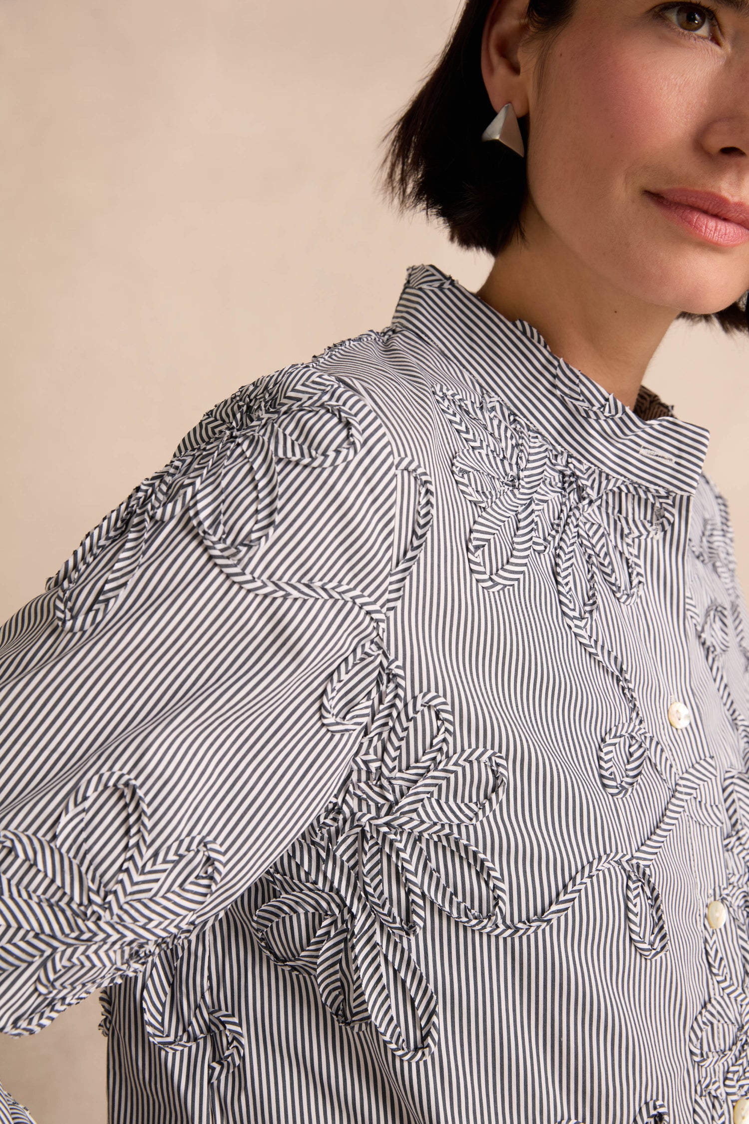 A close-up shows a woman wearing the Embroidered Stripe Shirt—a cotton-rich black and white striped top with embroidered floral details—paired with geometric earrings, photographed from the shoulders up against a neutral background.