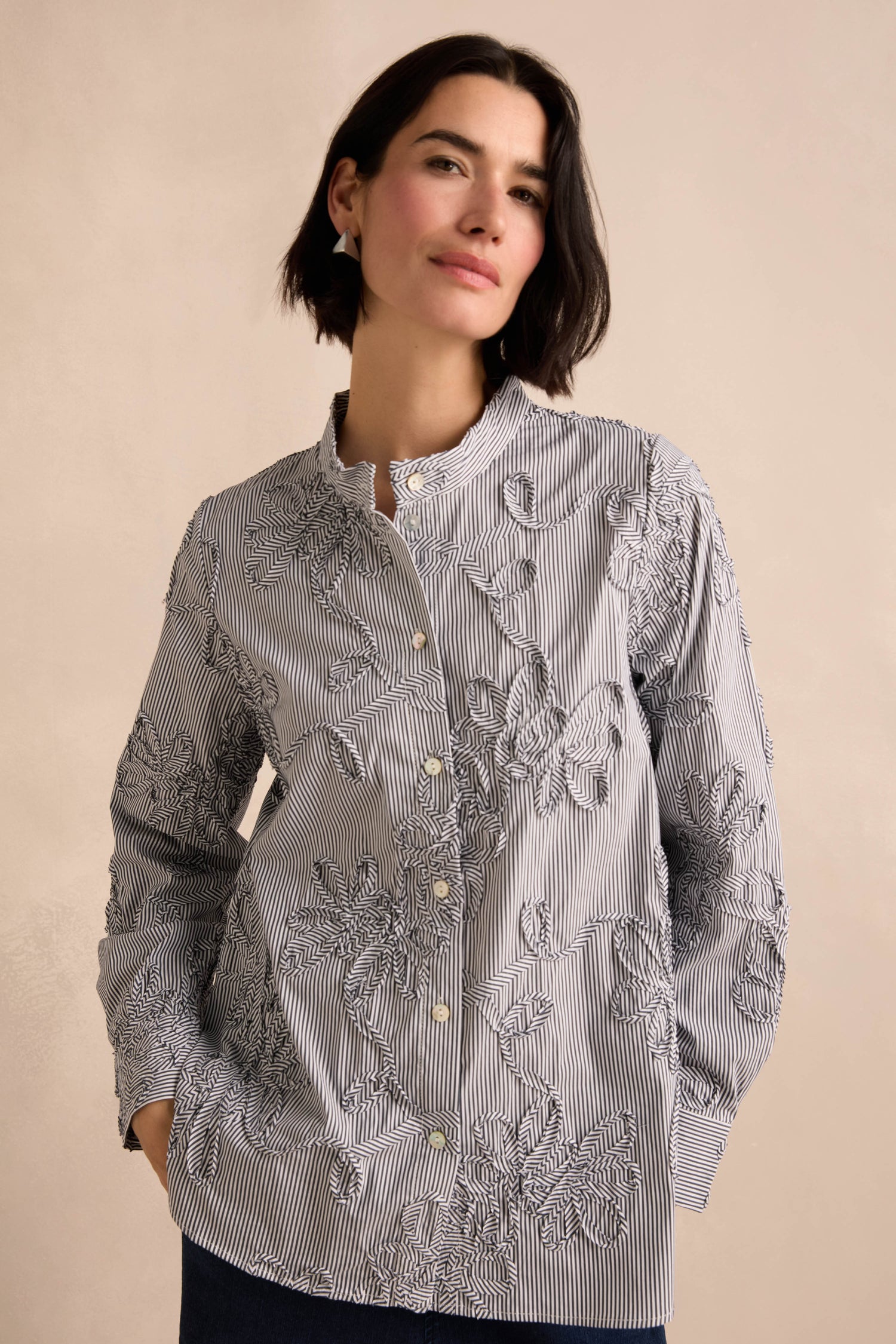 A woman wearing the Embroidered Stripe Shirt, featuring cotton-rich striped fabric and delicate floral embroidery, stands with one hand in her pocket against a neutral background.