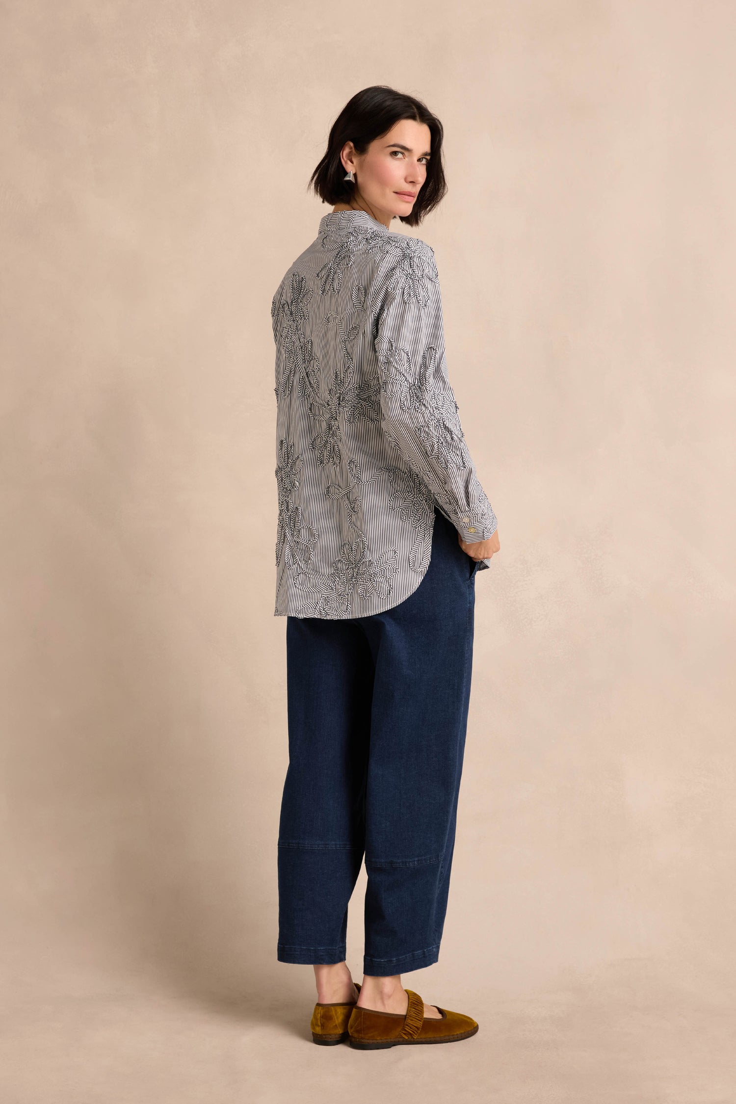 A woman with short dark hair wears the Embroidered Stripe Shirt—a textured gray, cotton-rich, long-sleeve top—paired with dark blue cuffed pants and brown shoes as she stands against a beige background, looking over her shoulder.