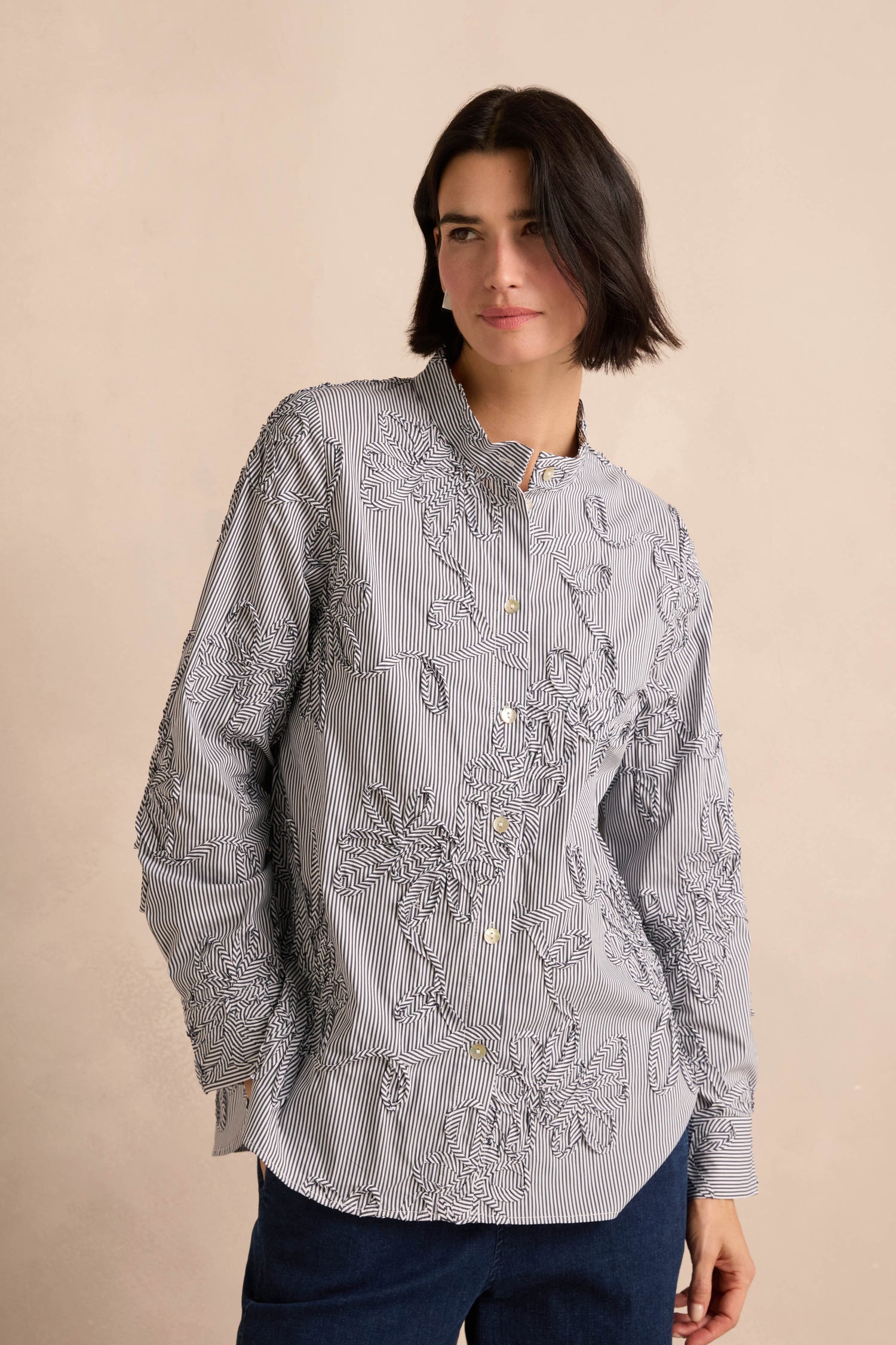 Wearing the Embroidered Stripe Shirt—a cotton-rich, long-sleeve, button-up top featuring stripe patterns and floral embroidery—a person with short dark hair pairs it with dark pants and stands against a beige background.