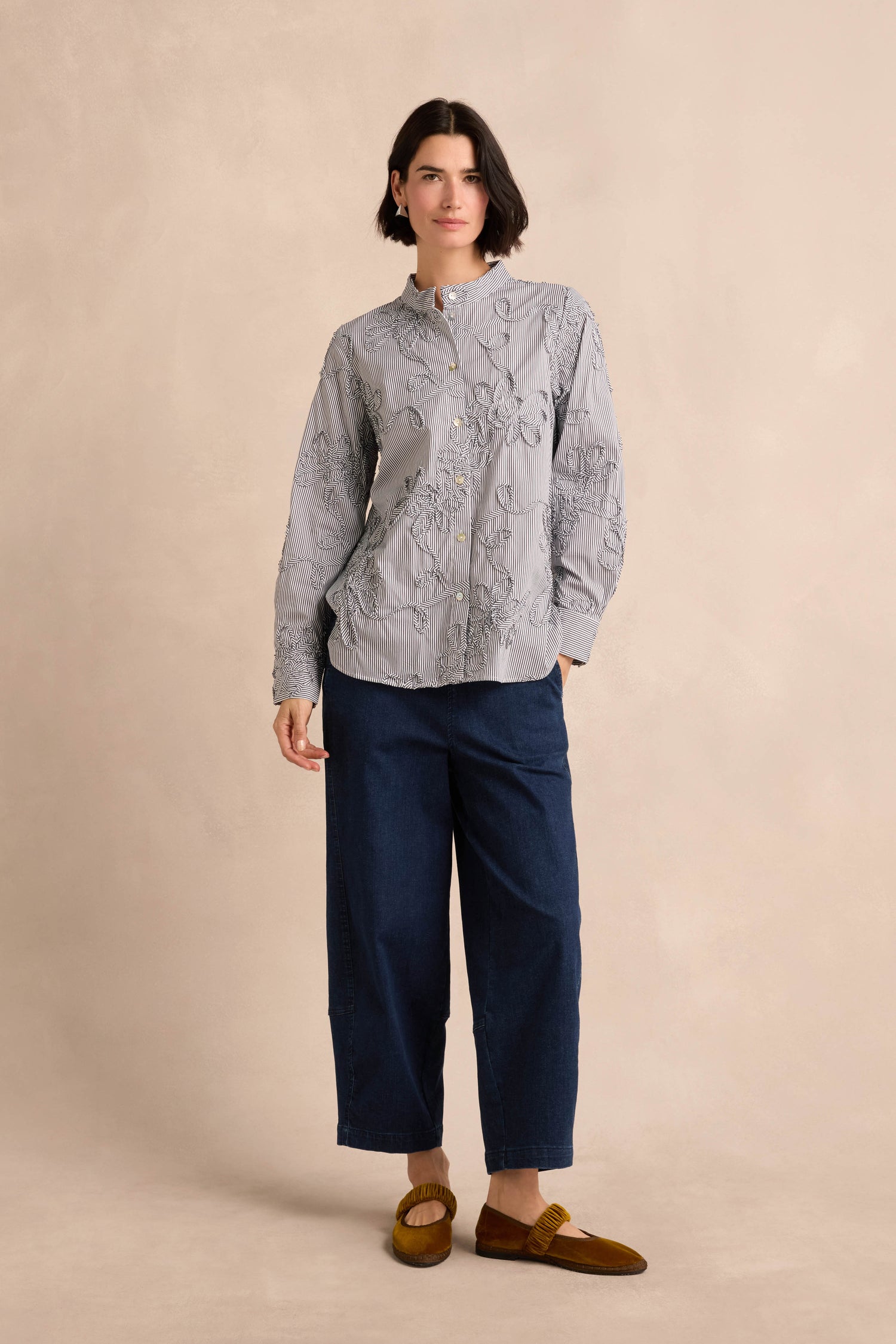A person stands against a beige background wearing the Embroidered Stripe Shirt, paired with dark blue wide-leg pants and brown loafers.