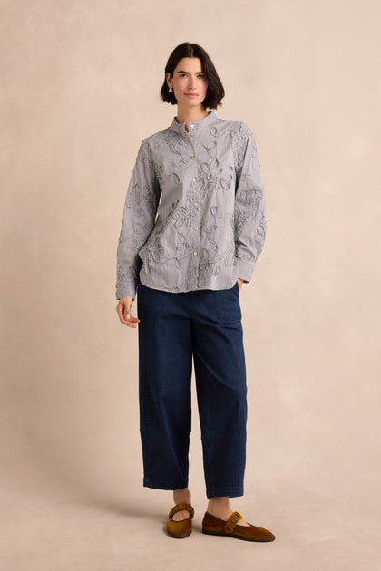 A person stands against a beige background wearing the Embroidered Stripe Shirt, paired with dark blue wide-leg pants and brown loafers.