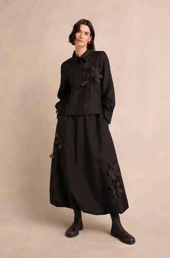 A woman wearing the Flower Detail Shirt—a loose black shirt with large floral appliqués—styled under a black jacket and paired with a long black skirt and black boots, stands against a plain beige background.