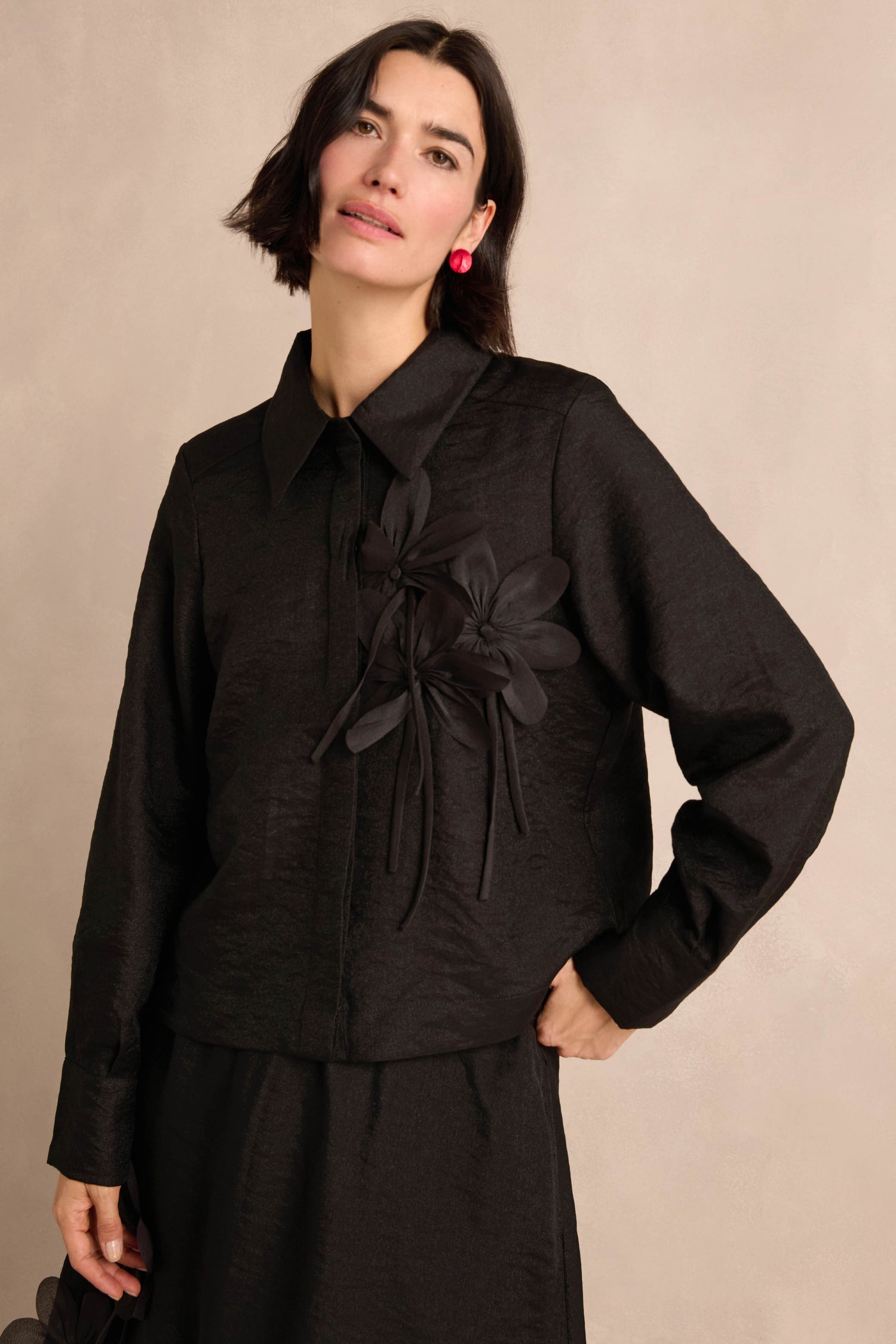A woman poses against a neutral background wearing the Flower Detail Shirt—a black textured jacket with floral embellishments on the chest—paired with a black skirt and red earrings.