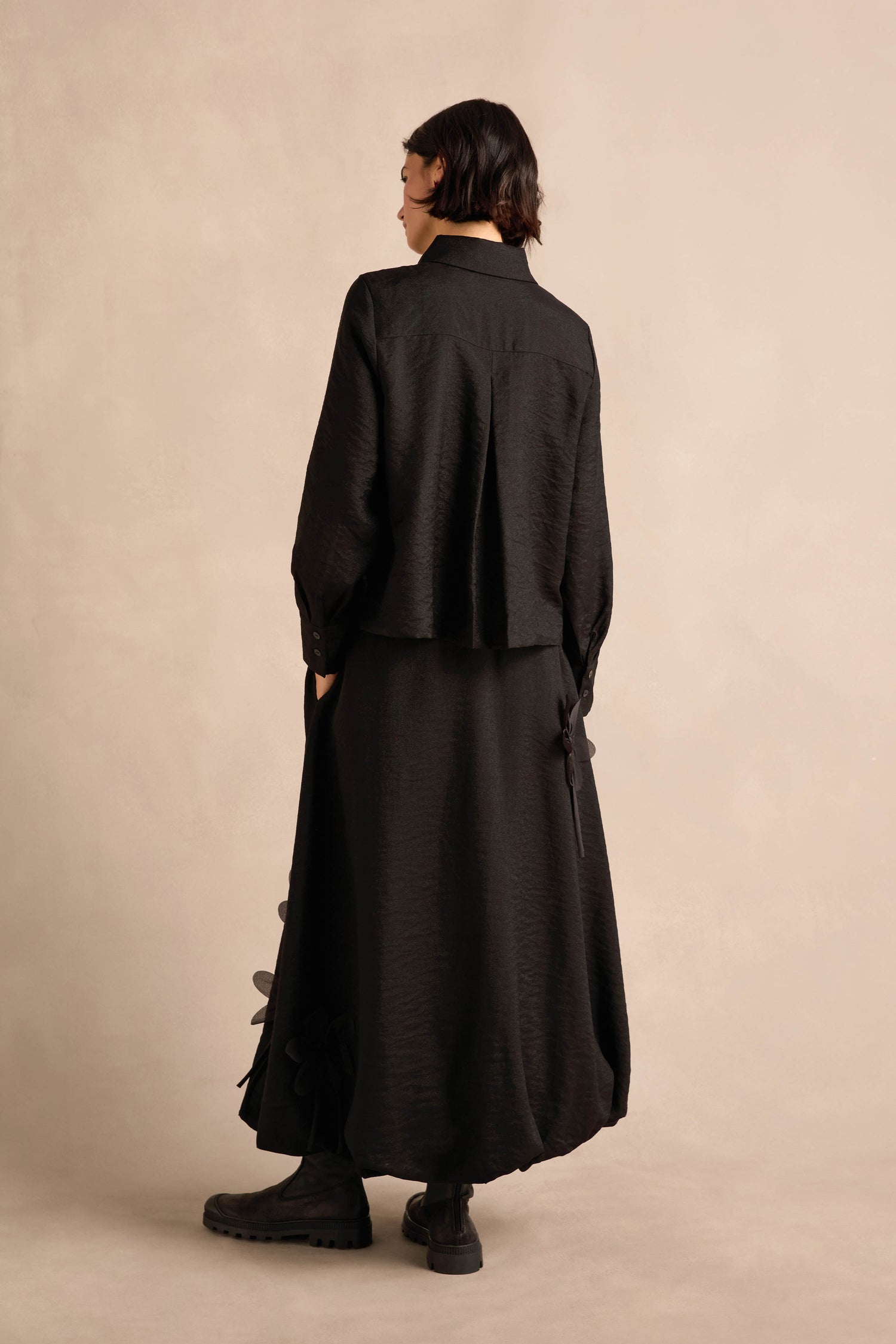 Person standing with back facing the camera, wearing the Flower Detail Shirt—a black shirt with floral appliqué—finished with a long black skirt featuring flower details and black boots, set against a light neutral background.