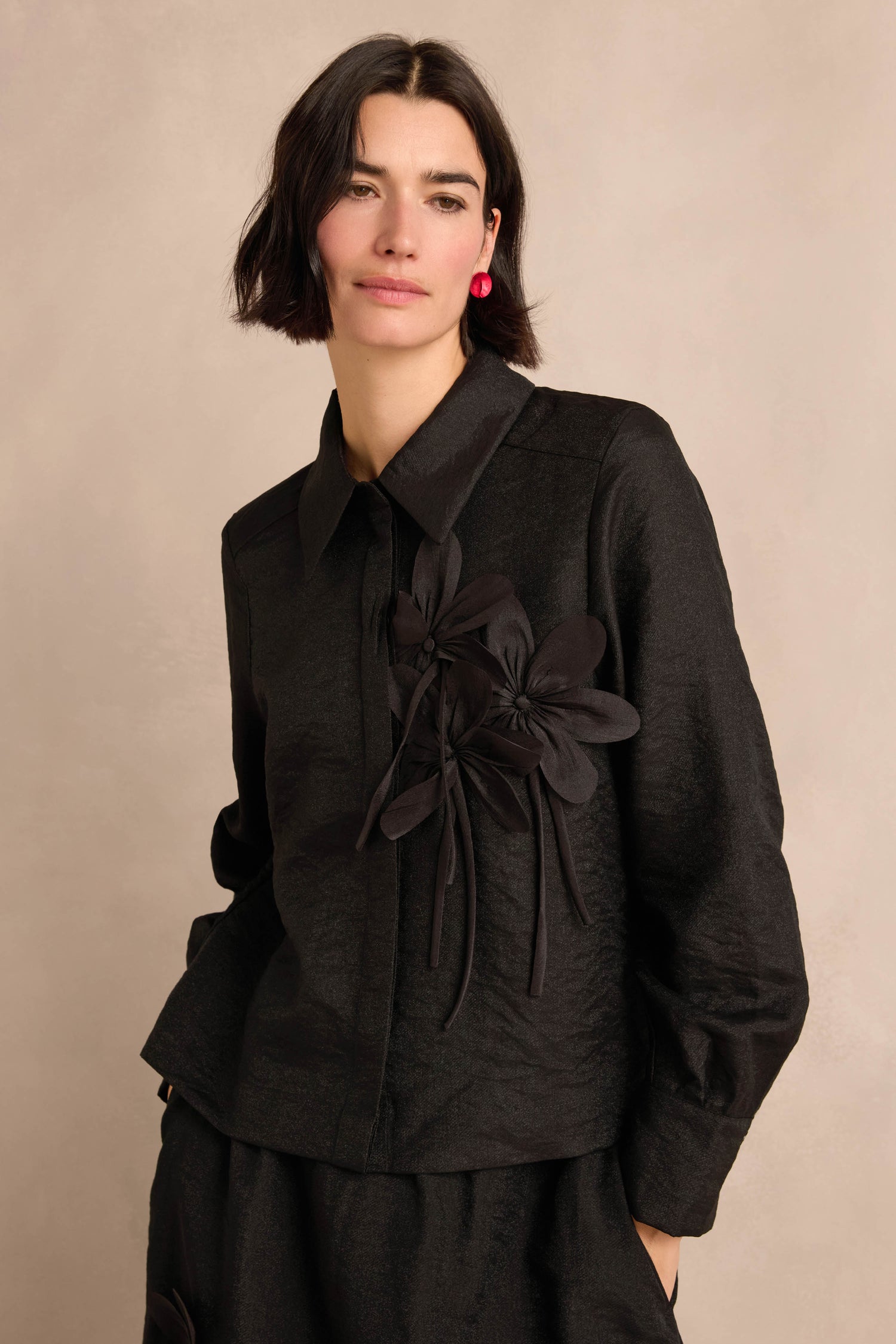 Woman with short dark hair wearing the Flower Detail Shirt—a textured black jacket featuring two floral appliqué embellishments—posing against a neutral background.