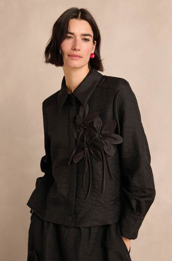 Woman with short dark hair wearing the Flower Detail Shirt—a textured black jacket featuring two floral appliqué embellishments—posing against a neutral background.