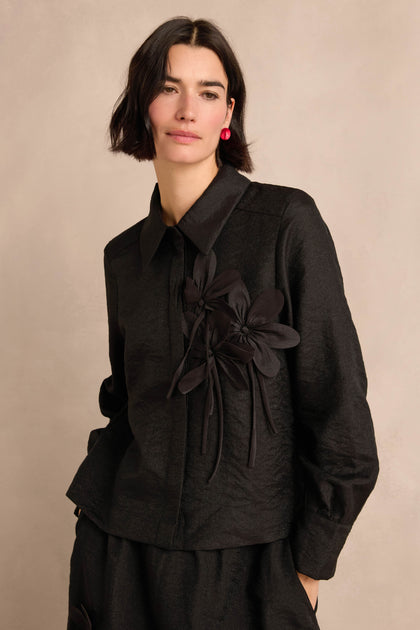 Woman with short dark hair wearing the Flower Detail Shirt—a textured black jacket featuring two floral appliqué embellishments—posing against a neutral background.