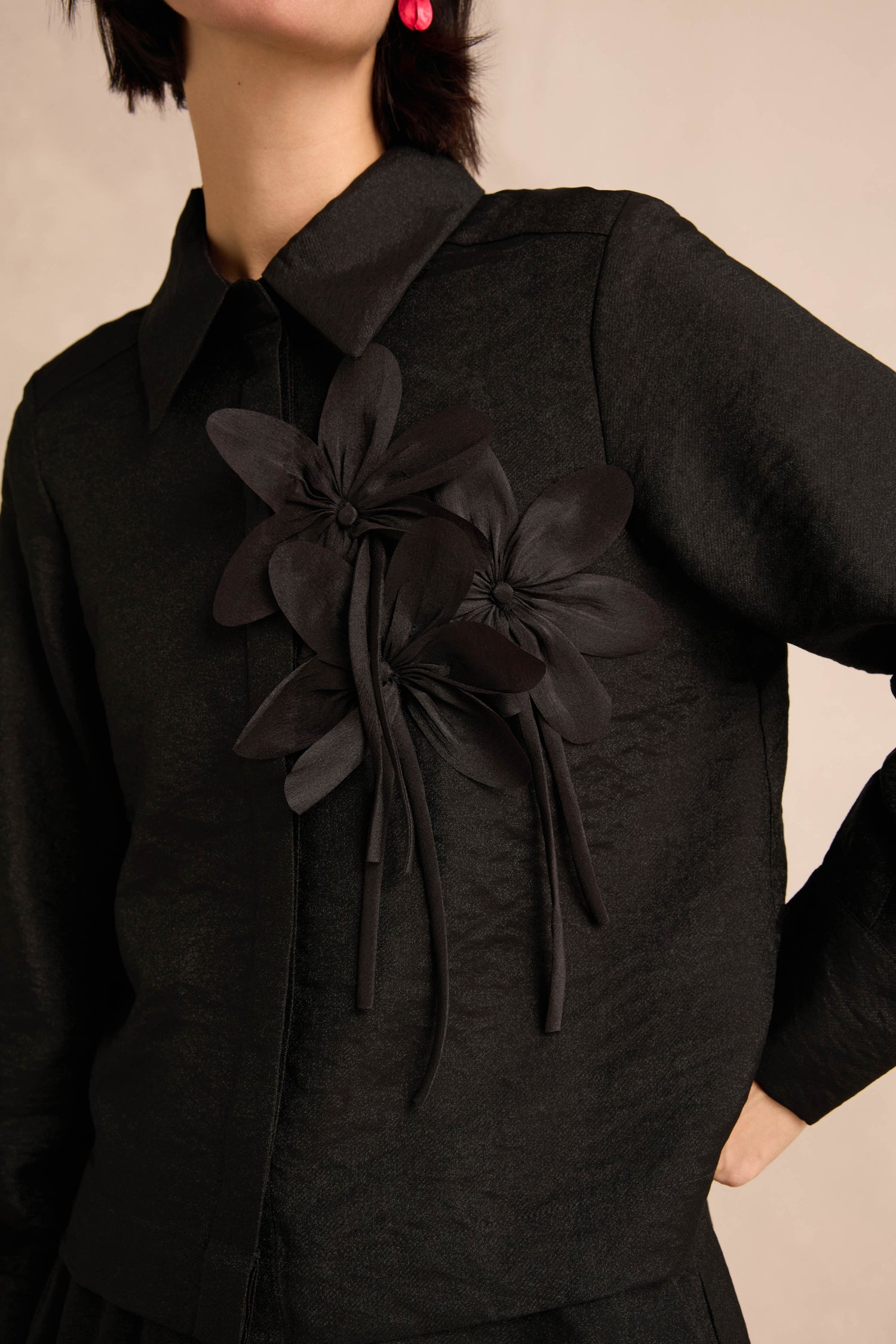A person wearing the Flower Detail Shirt—a textured black top featuring two large floral appliqué embellishments on the chest—stands against a neutral background.