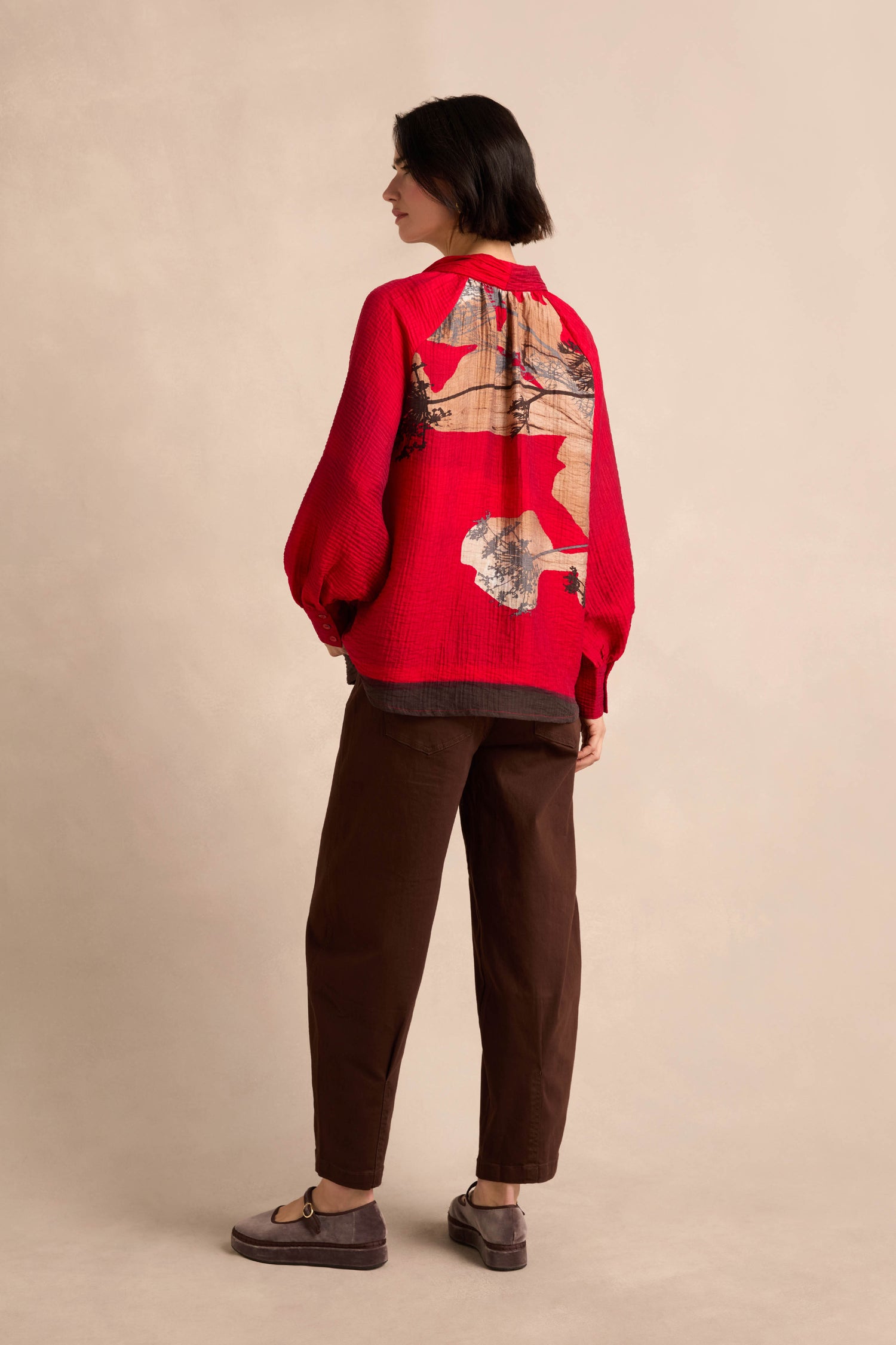 A person stands sideways against a beige background, wearing the Kyoto Puff Sleeve Shirt in a red patterned textured fabric, paired with brown pants and loafers.