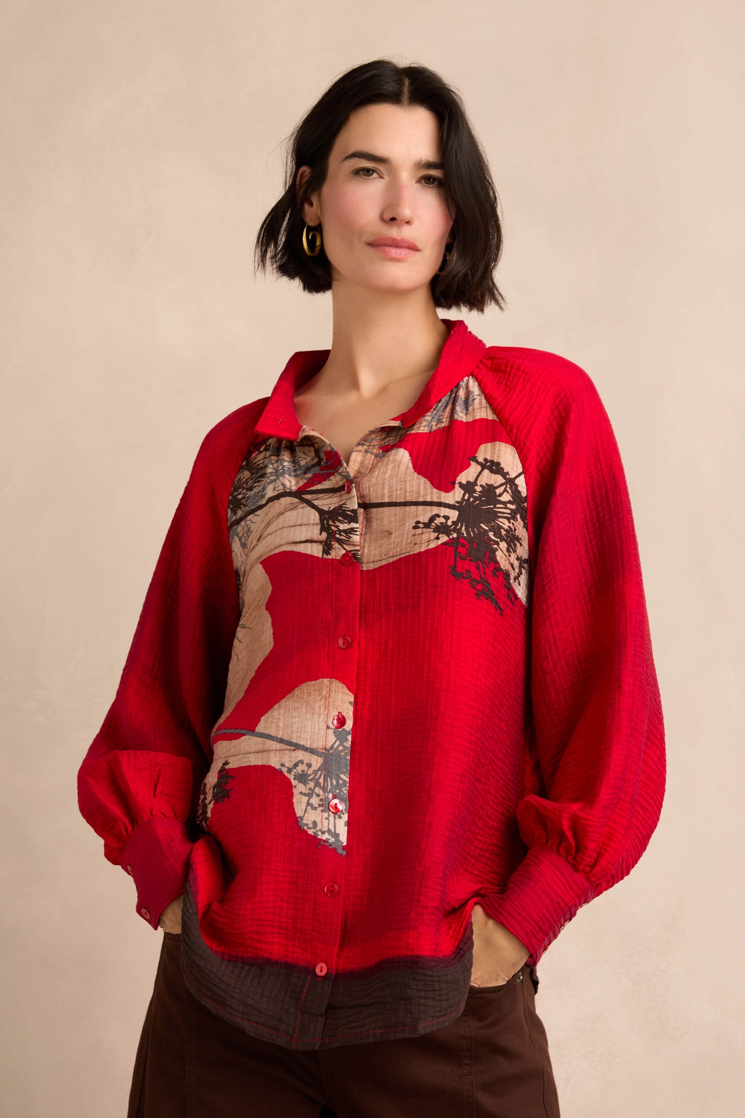 A woman wears the Kyoto Puff Sleeve Shirt, featuring a red hue with beige and brown abstract placement print patterns, as she stands with her hands in her pockets against a neutral background.