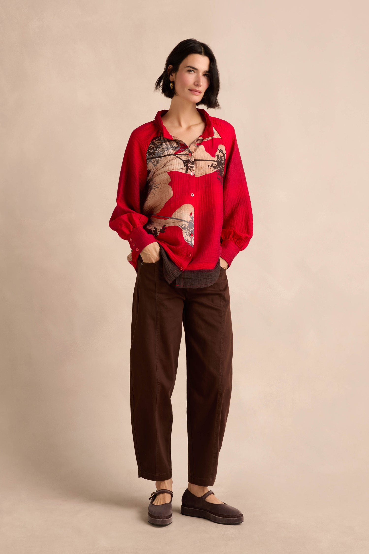 A woman stands against a neutral background, hands in pockets, wearing the Kyoto Puff Sleeve Shirt—crafted from textured fabric with a red placement print—paired with brown pants and matching shoes.