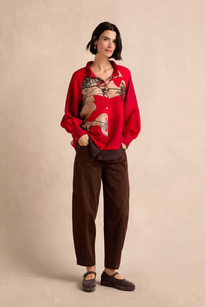 A woman stands against a neutral background, hands in pockets, wearing the Kyoto Puff Sleeve Shirt—crafted from textured fabric with a red placement print—paired with brown pants and matching shoes.