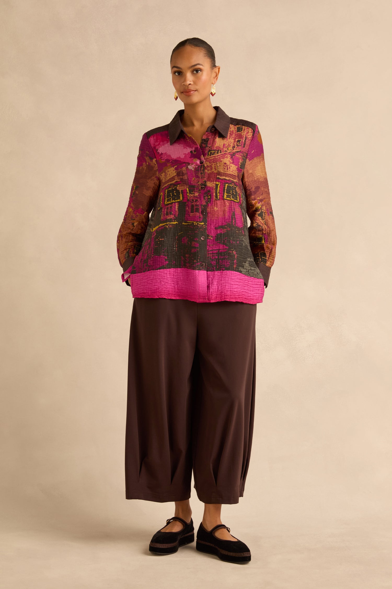 A person wearing a Window Print Pleated Shirt with an abstract architectural print and wide-leg dark brown trousers.