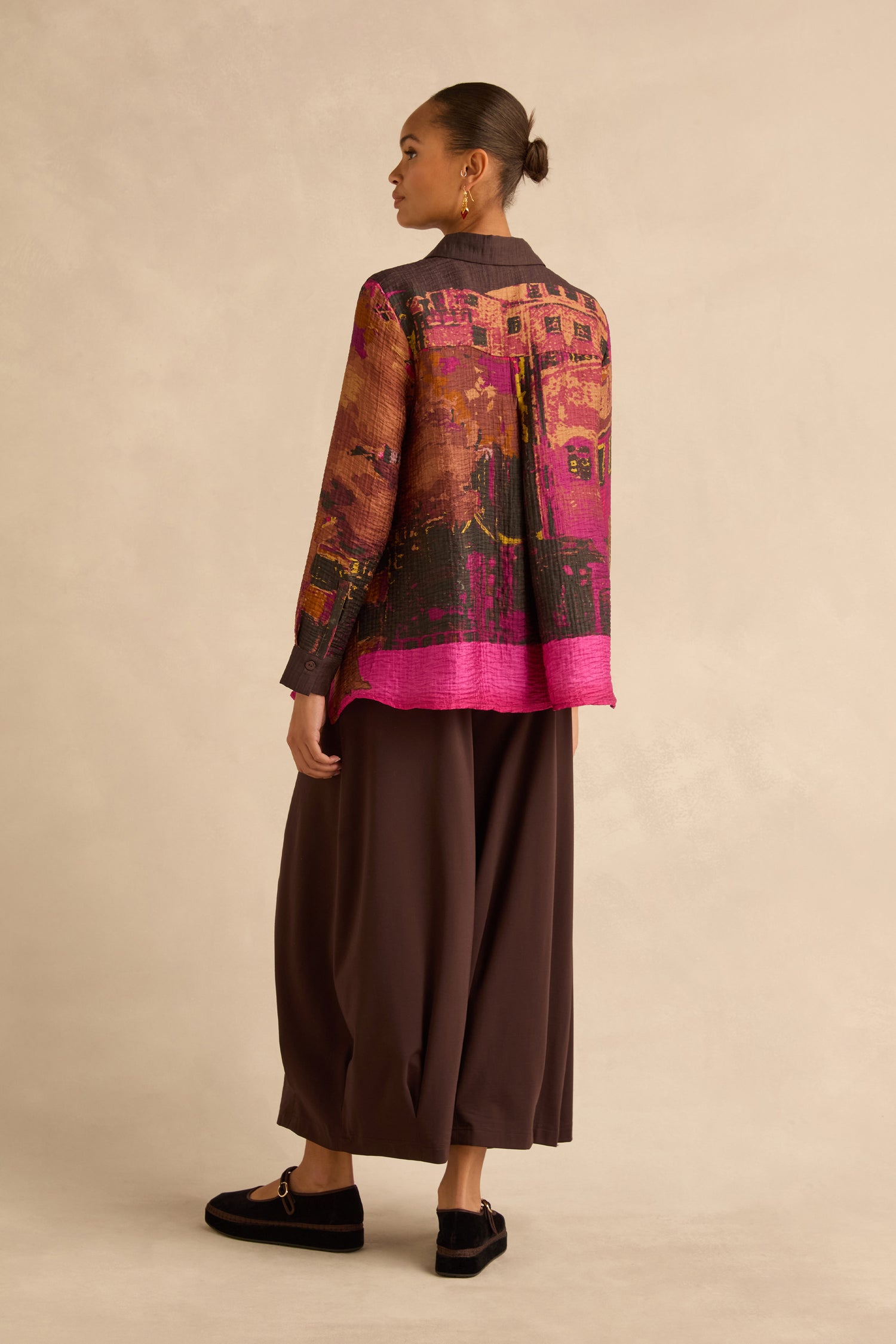 A person viewed from behind wearing a Window Print Pleated Shirt with an abstract architectural print and wide-legged dark brown trousers.