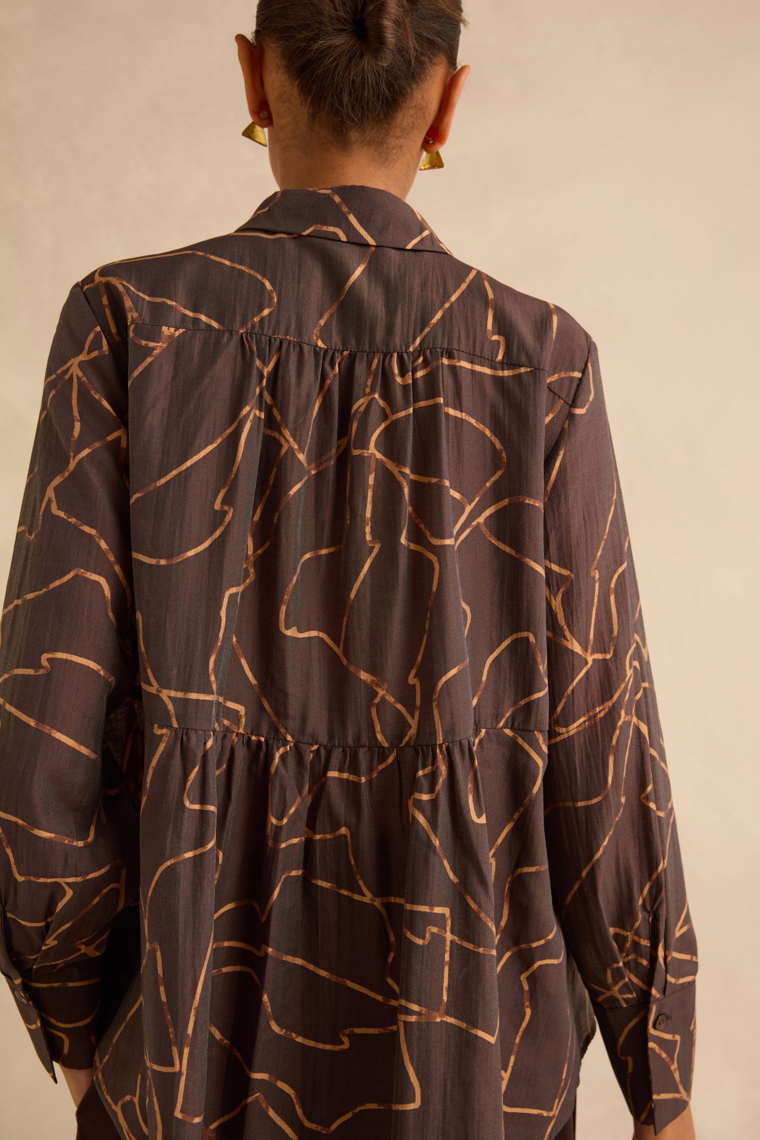 Back view of a person wearing a Swirl Print Sheer Shirt in dark brown with a gold line-art swirl print and a gathered tier at the waist.