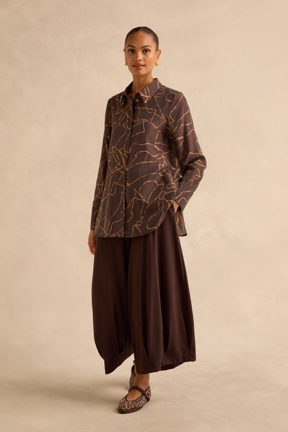 A person wearing a Swirl Print Sheer Shirt in brown with a gold line print, paired with a dark brown, wide-leg, ankle-length skirt and leopard-print flats.