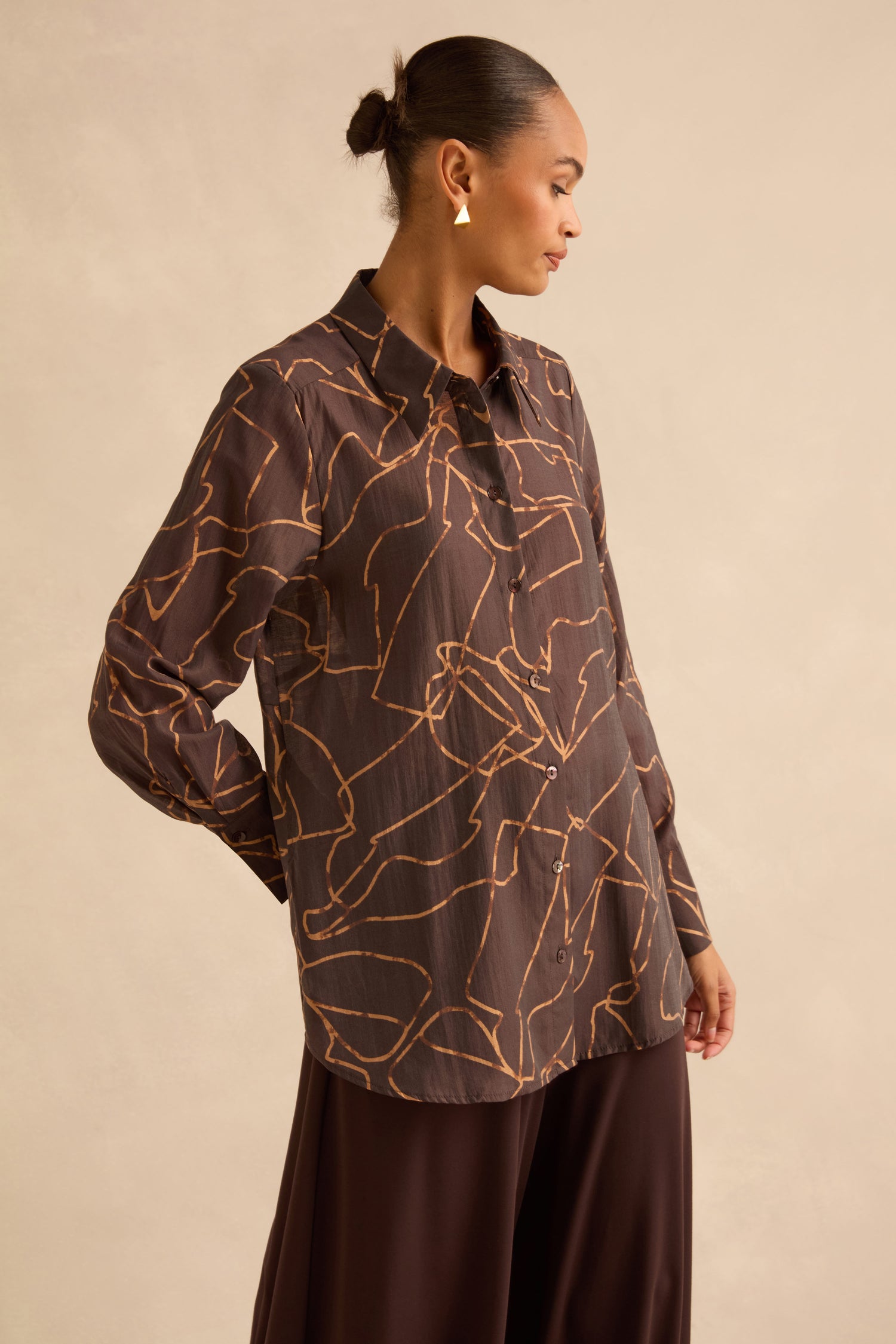 A person wearing a brown Swirl Print Sheer Shirt with a sheer, long-sleeved button-up design and an abstract gold line print.