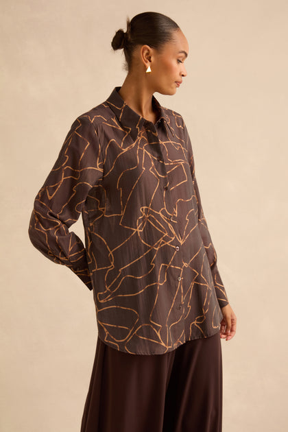 A person wearing a brown Swirl Print Sheer Shirt with a sheer, long-sleeved button-up design and an abstract gold line print.