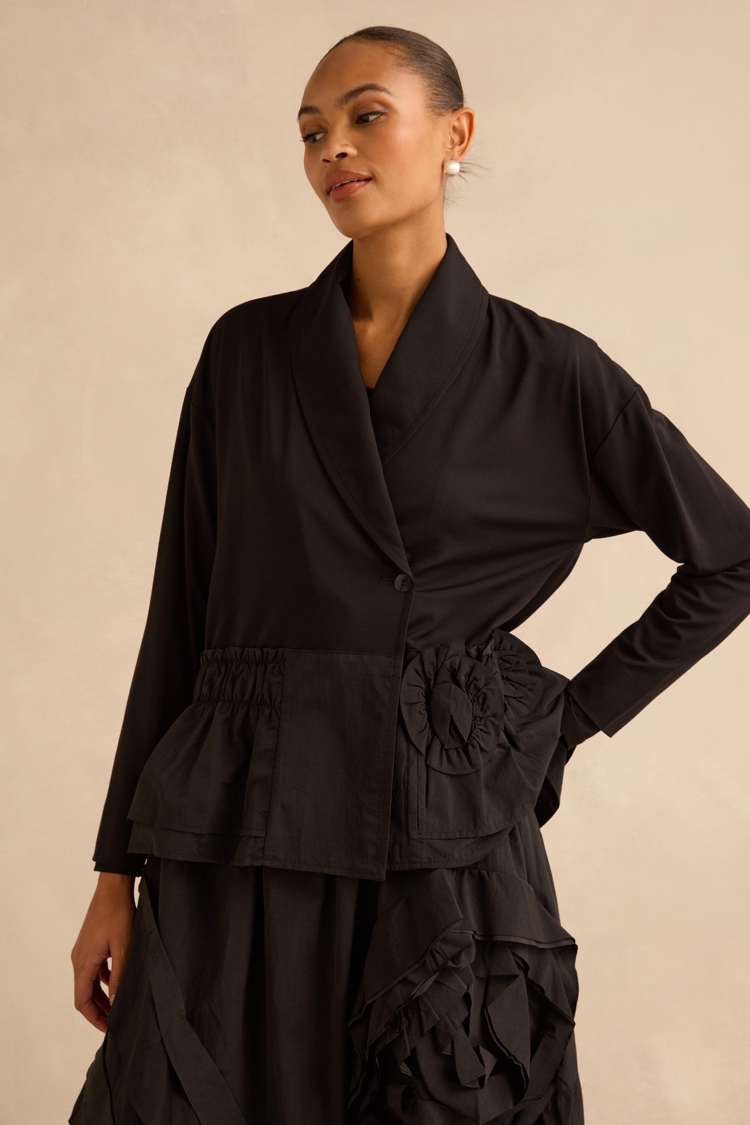 A person wears a black Taffeta Frill Hem Jacket featuring a shawl collar, a gathered waist, and ruffled hem details.