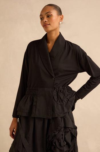 A person wears a black Taffeta Frill Hem Jacket featuring a shawl collar, a gathered waist, and ruffled hem details.