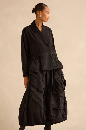 A woman wearing a black Taffeta Frill Hem Jacket with a shawl collar, drop-waist seam, and a ruffled, layered skirt.