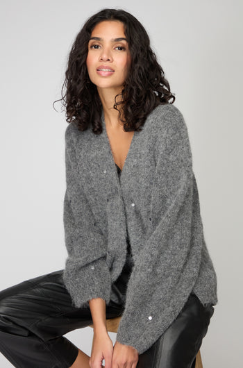 A woman with curly dark hair sits on a stool, wearing the Sequin Cardi and black leather pants, capturing Nordic minimalism against a plain light background.