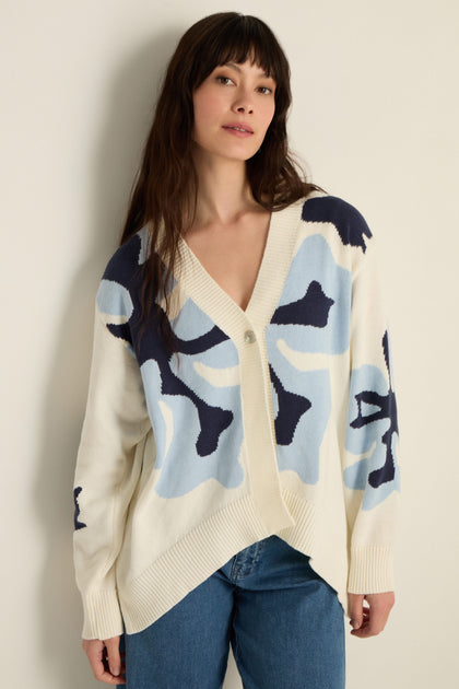 A woman in the Ginkgo Knit Cardigan, featuring a relaxed fit and abstract blue patterns on a cotton blend, layered over a white top and blue jeans, stands against a plain light wall.