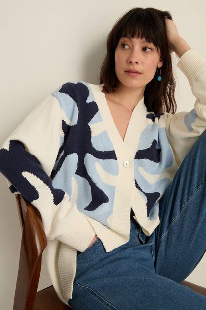 A woman sits on a chair wearing the Ginkgo Knit Cardigan—a cream cotton-blend with blue abstract patterns—and blue jeans, looking to the side.