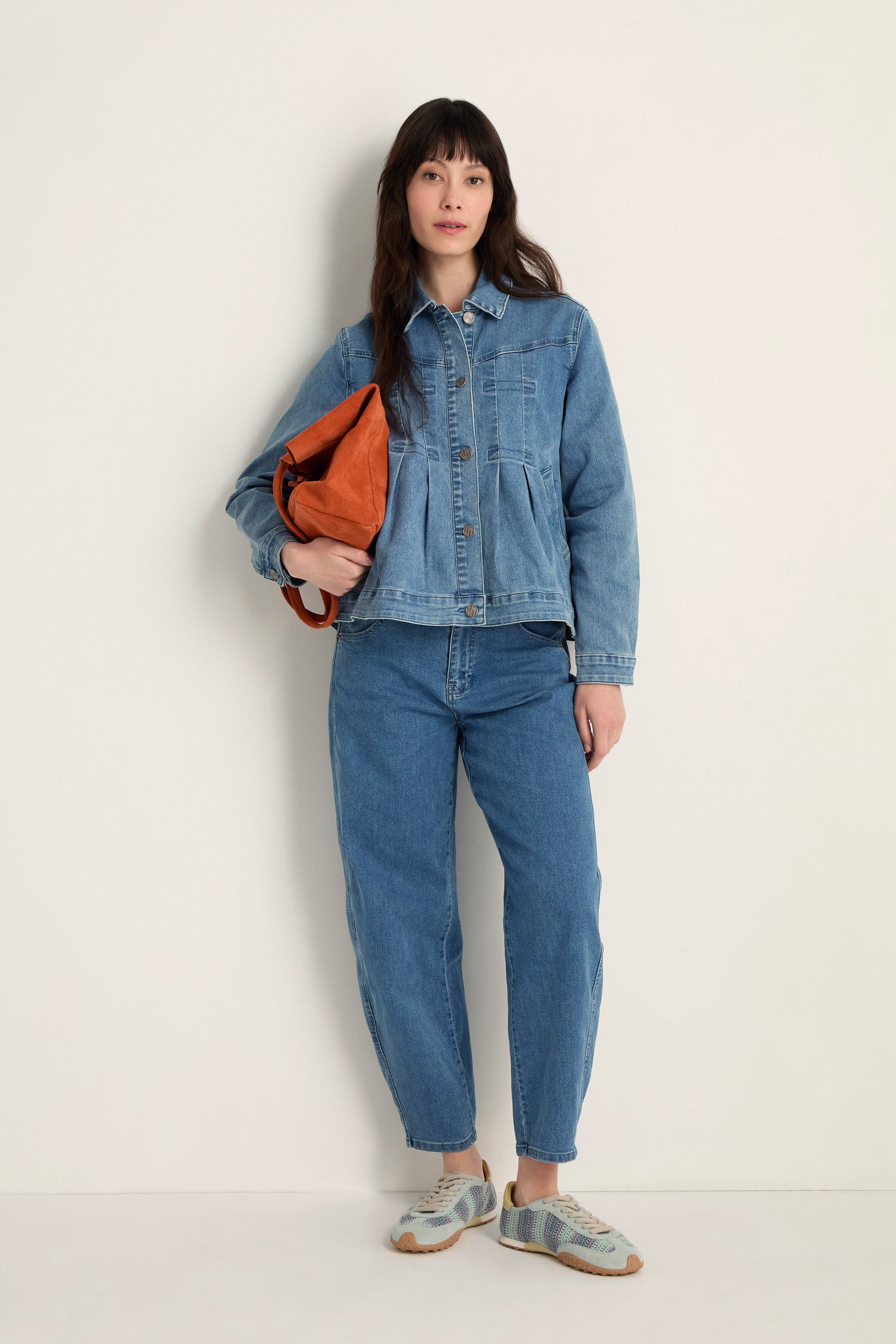 A woman stands against a plain wall wearing the Cropped Denim Jacket in stretch cotton denim, styled with blue jeans, patterned sneakers, and an orange bag—a sleek, contemporary layer for any casual look.