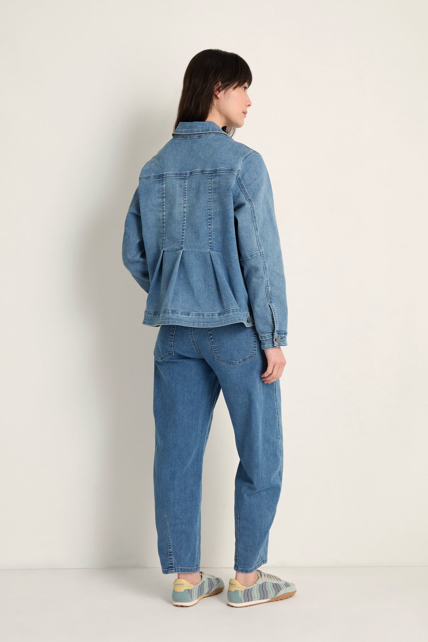 A person faces away, wearing the Cropped Denim Jacket in light blue stretch cotton denim with matching jeans and multicolored sneakers, set against a plain light background for a modern, contemporary look.