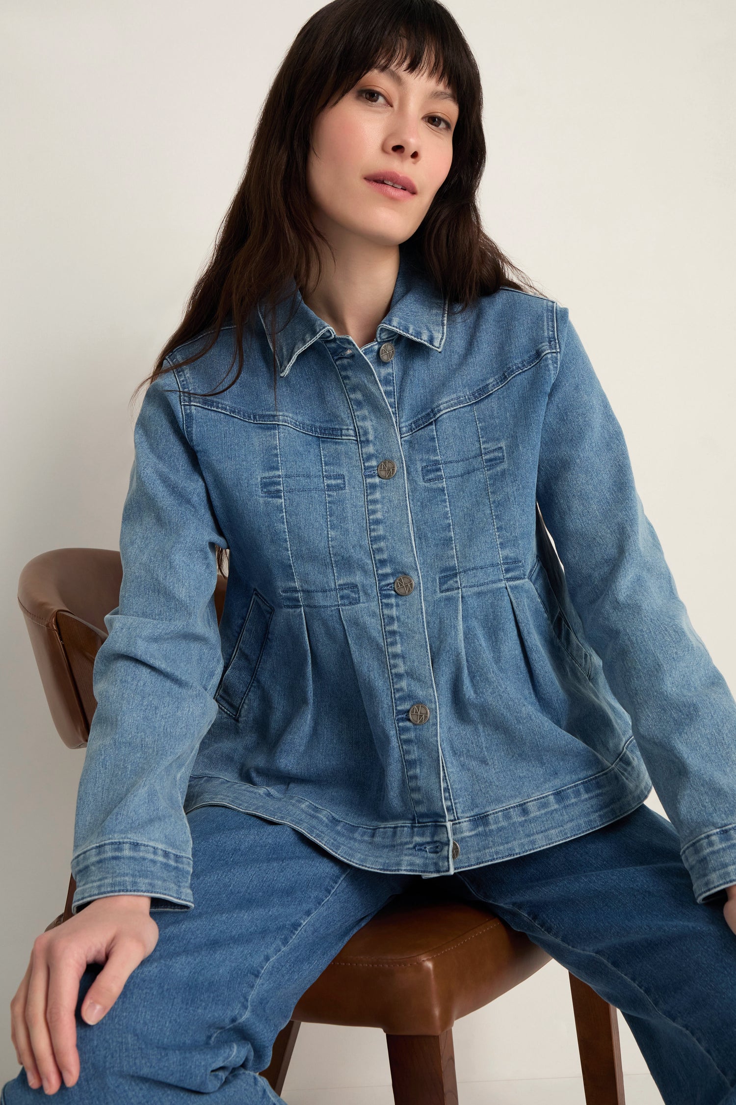 A woman with long dark hair sits on a chair wearing the Cropped Denim Jacket and matching jeans in stretch cotton denim, creating a modern look against a plain background.