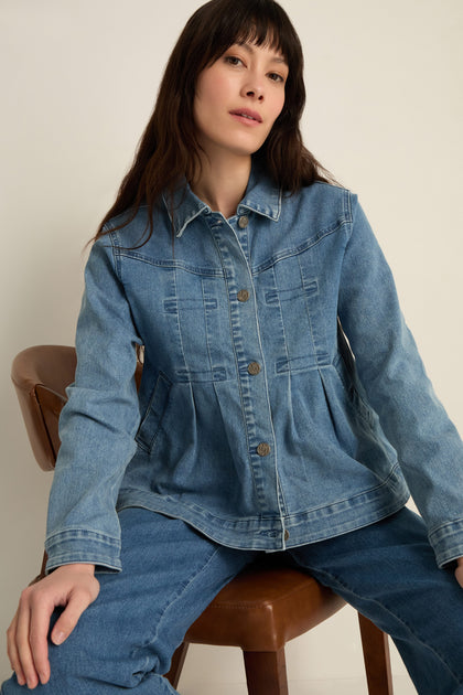 A woman with long dark hair sits on a chair wearing the Cropped Denim Jacket and matching jeans in stretch cotton denim, creating a modern look against a plain background.