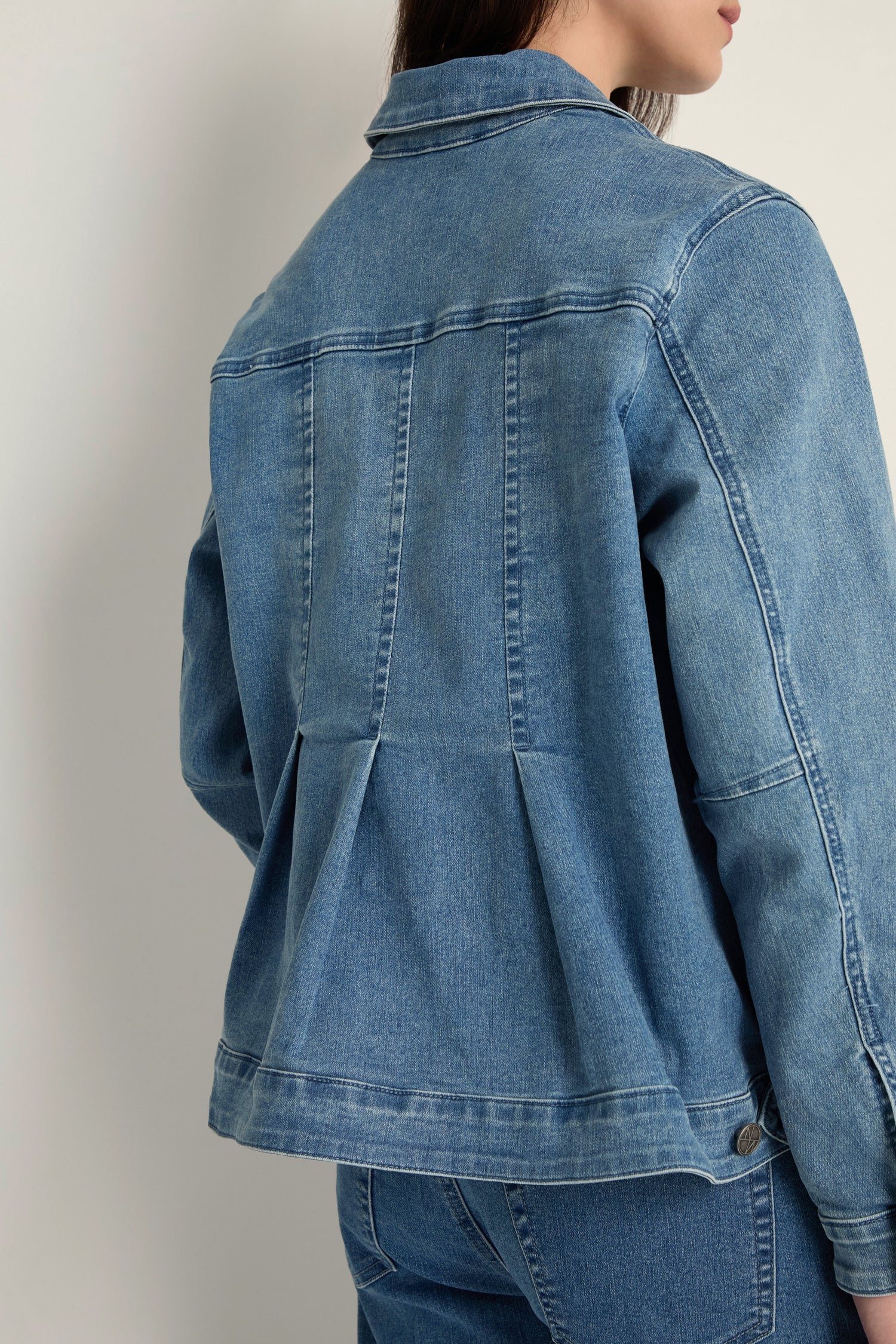 A person wears the Cropped Denim Jacket featuring pleated back details, paired with matching stretch cotton blue denim jeans, standing against a plain light background for a contemporary, effortless look.
