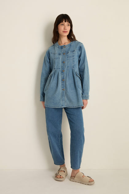 A woman stands against a plain wall wearing the Long Denim Jacket, a modern layering piece made from soft stretch denim, styled with blue jeans and beige sandals.