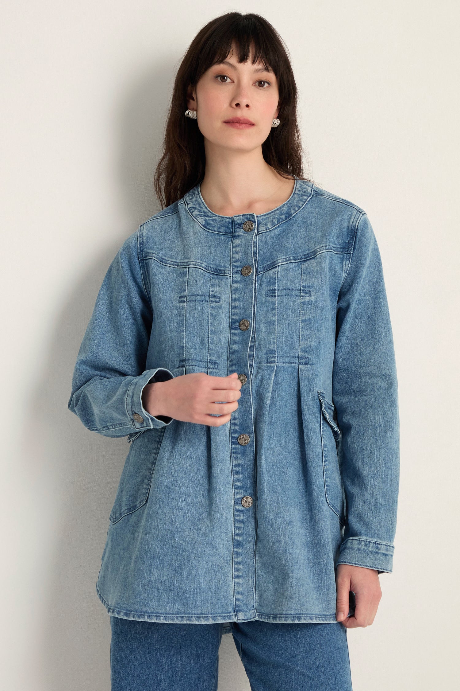 A woman stands against a plain light wall wearing the Long Denim Jacket, made from soft stretch denim, and matching jeans—an effortlessly chic contemporary layering piece.