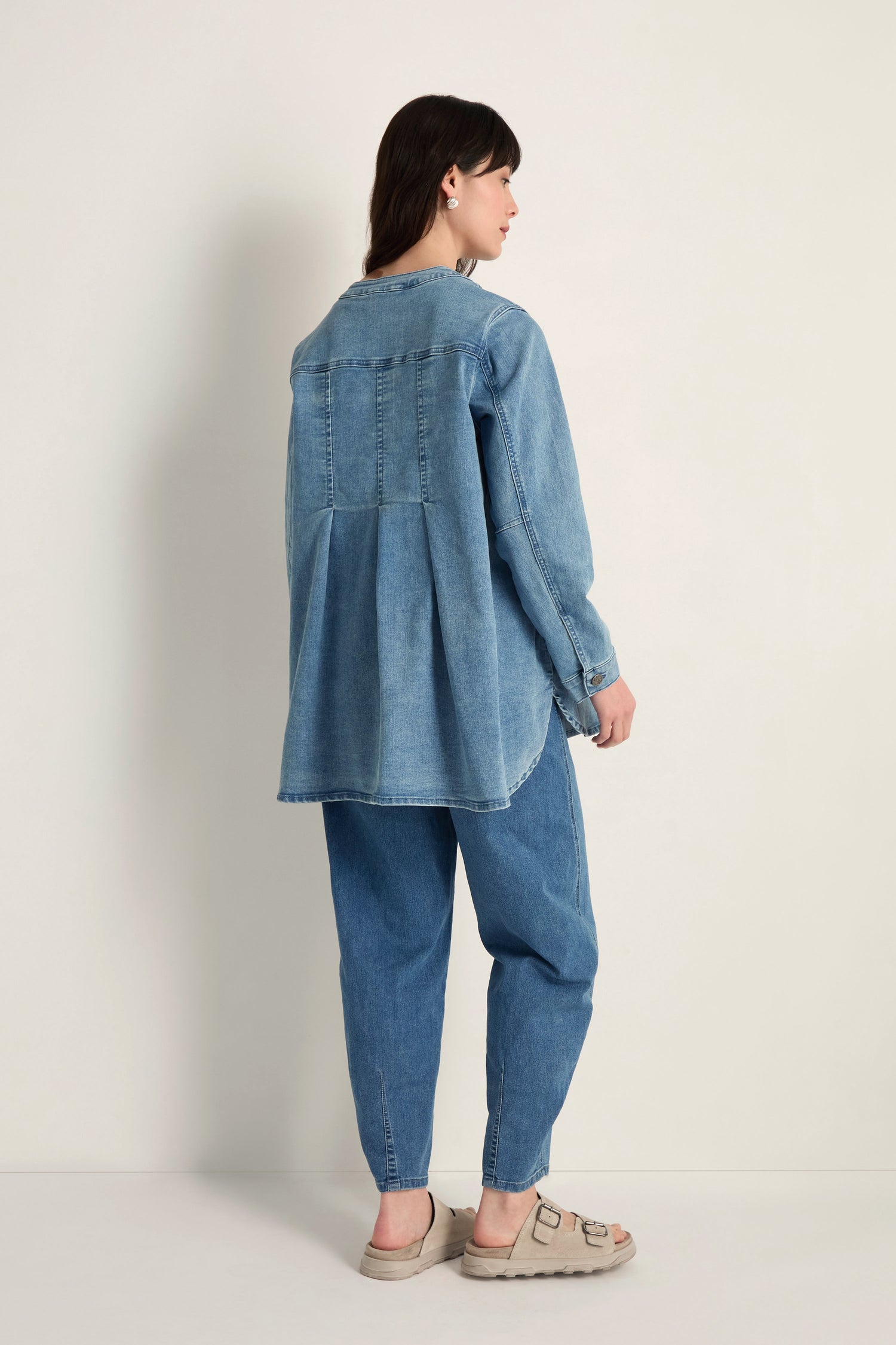 Someone faces away, wearing the Long Denim Jacket made from soft stretch denim and blue jeans, paired with beige sandals against a plain light background.