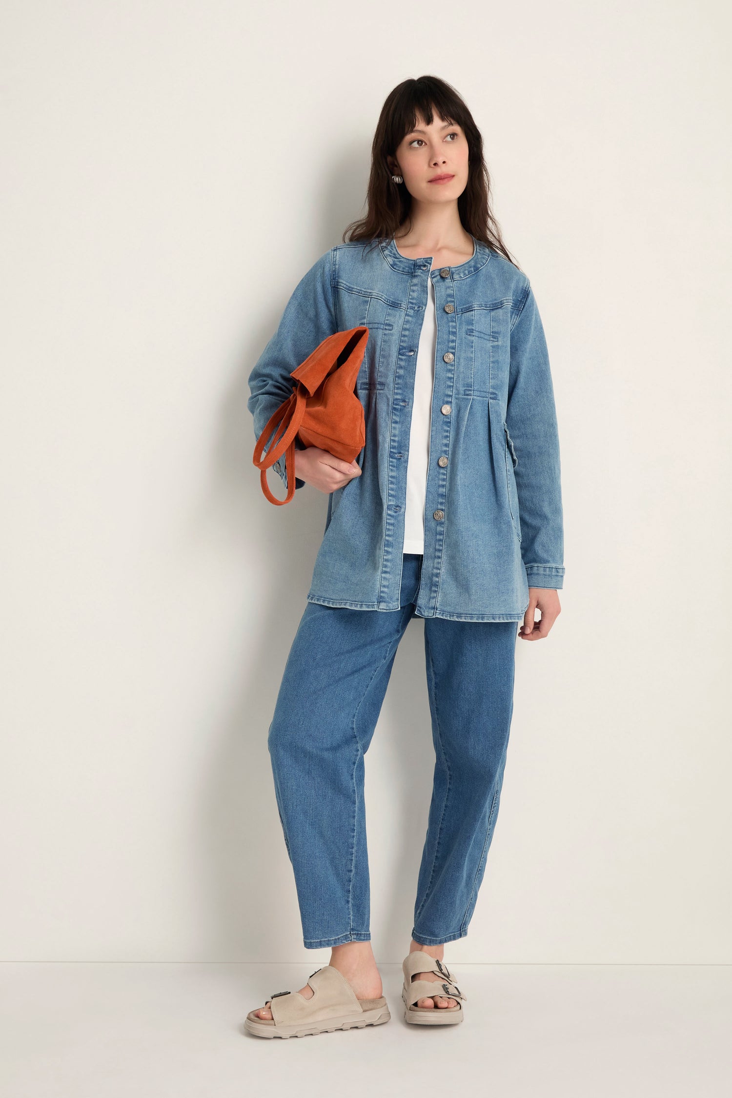 A woman stands against a plain wall wearing the Long Denim Jacket made from soft stretch denim with jeans, holding a brown bag and wearing beige sandals—a perfect contemporary layering piece.