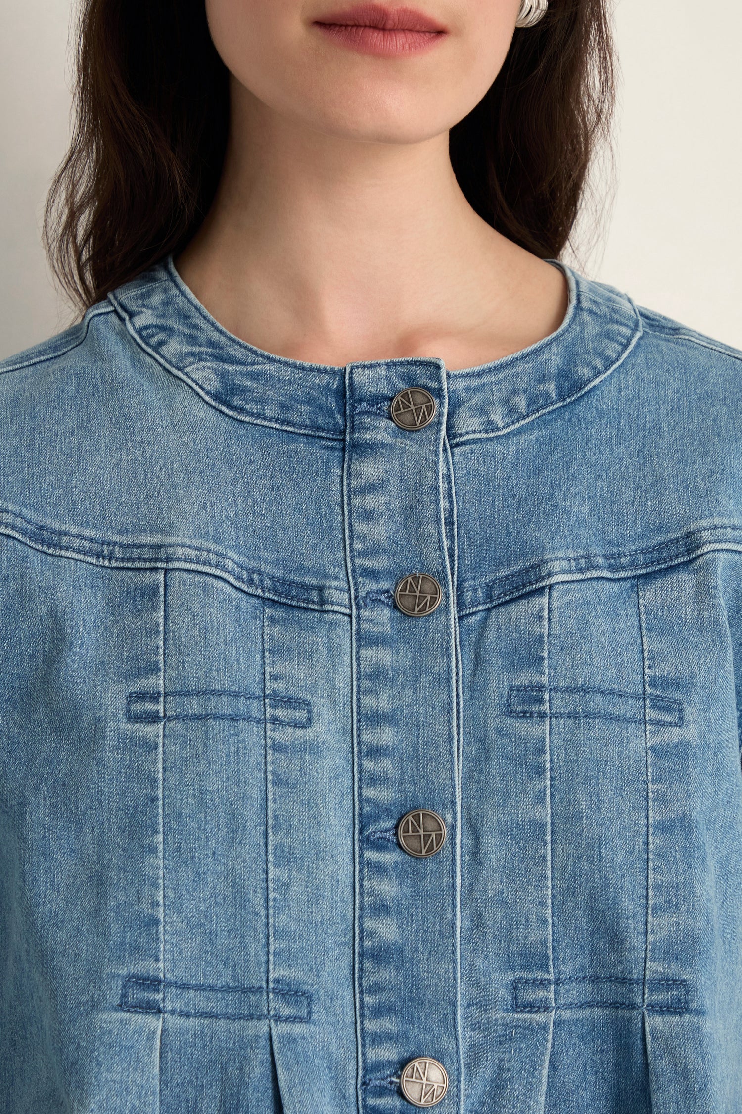 A woman wears the Long Denim Jacket, a round-neck, button-up top crafted from soft stretch denim with visible seams and metallic logo-embossed buttons.
