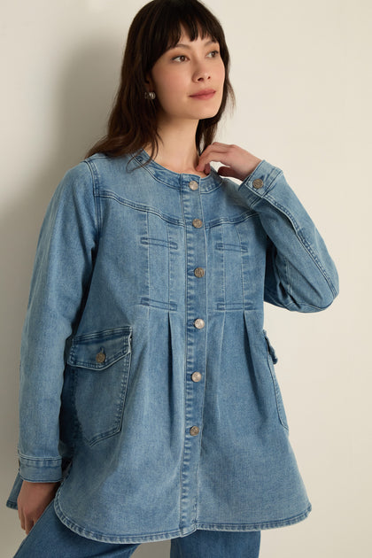 A woman stands against a plain wall wearing the Long Denim Jacket, a modern blue button-up with front pockets and long sleeves, made from soft stretch denim—perfect for layering.