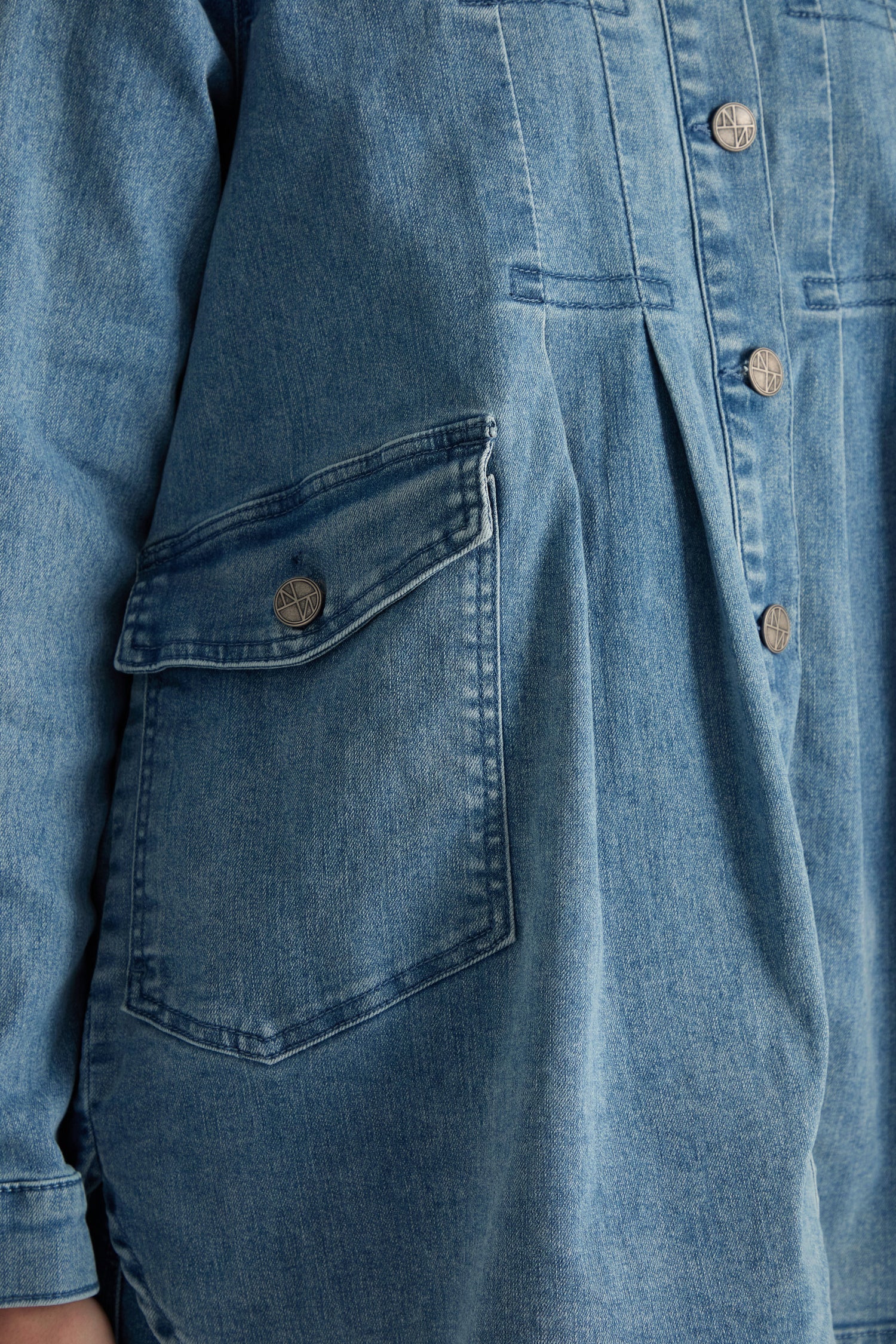 Close-up of the Long Denim Jacket in blue stretch denim, showcasing a large buttoned pocket, visible seams, metal buttons, and part of the left sleeve—a modern piece ideal for contemporary layering.