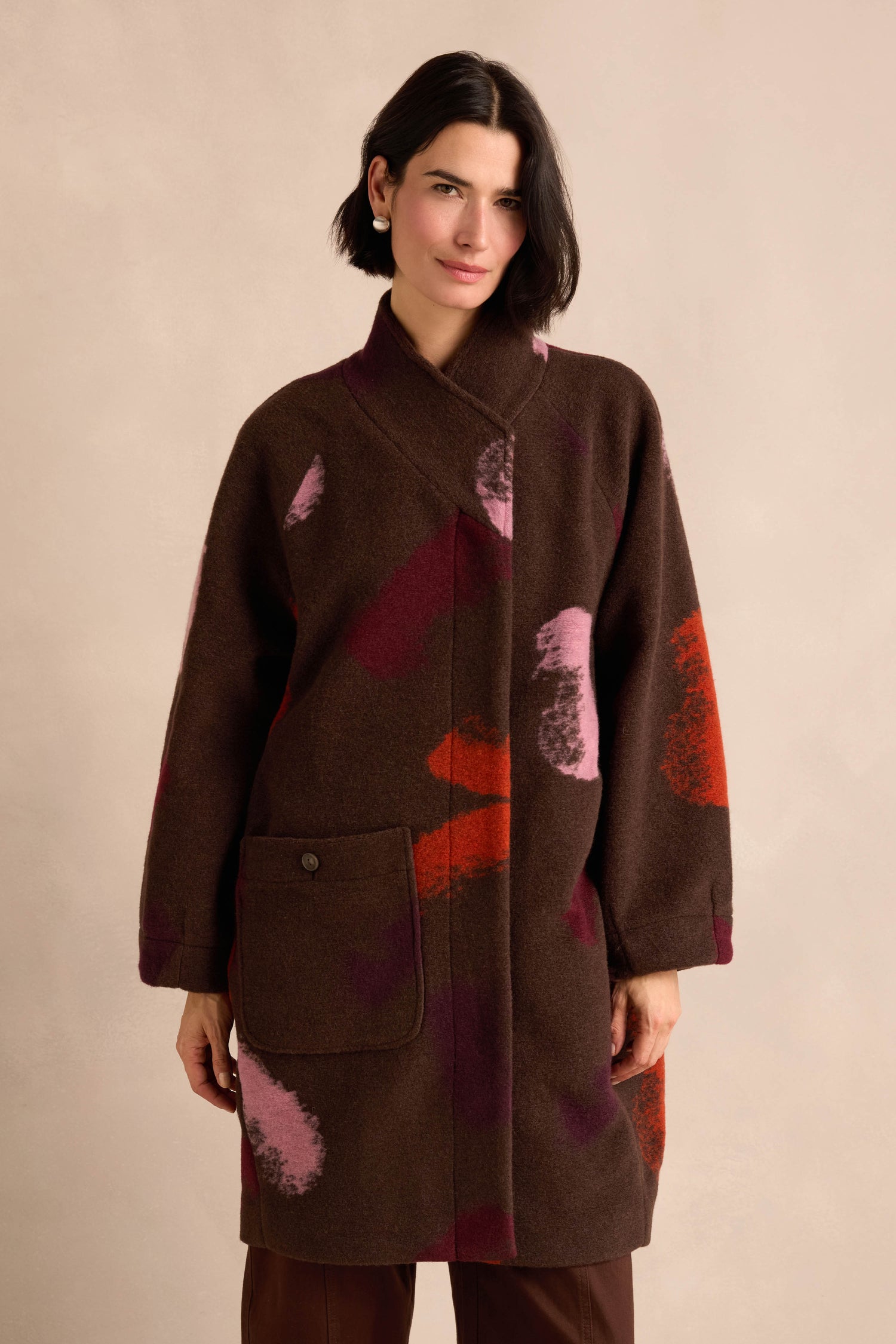 A woman wearing the Colour Splash Coat—a striking long winter coat crafted from a wool blend and adorned with large pink, red, and purple abstract patterns—stands against a beige background.