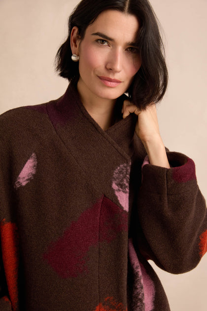 A woman with short dark hair wears the Colour Splash Coat—a long winter coat with a brown pattern highlighted by red and pink accents—looking at the camera with one hand touching her collar.