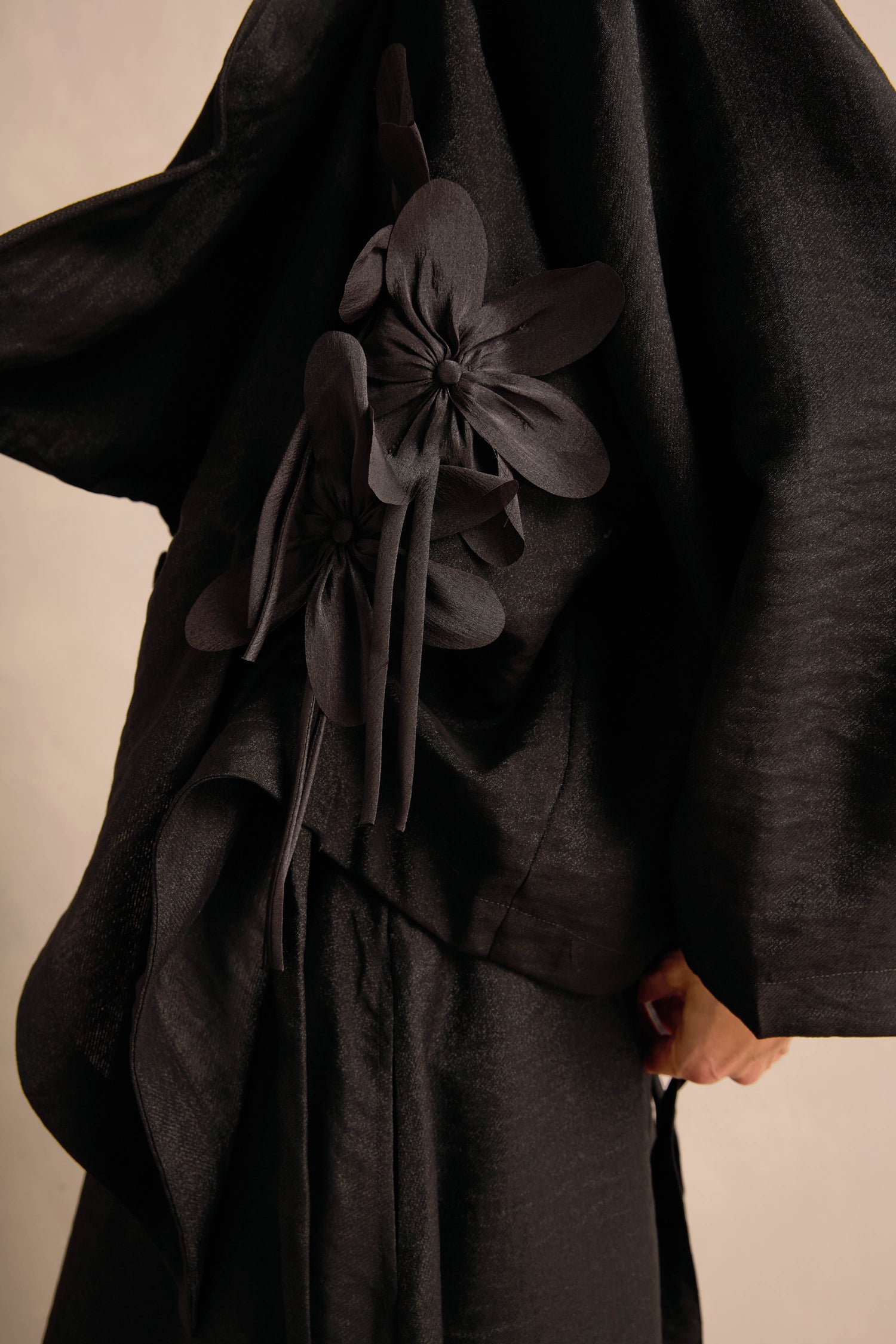 A person wearing the Flower Detail Jacket, a textured black draped jacket adorned with large fabric floral embellishments on the side.