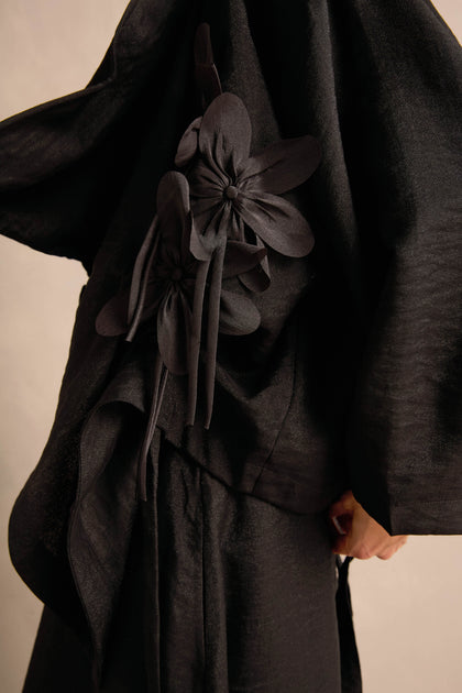 A person wearing the Flower Detail Jacket, a textured black draped jacket adorned with large fabric floral embellishments on the side.