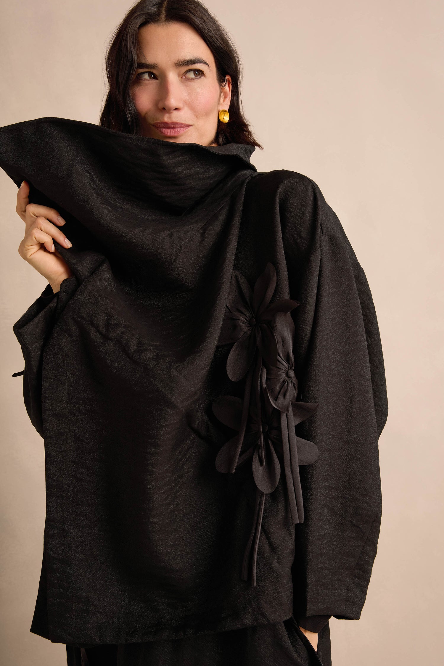 A woman wearing the Flower Detail Jacket—a textured black draped piece featuring an oversized collar and delicate floral appliqué—stands against a plain beige background.