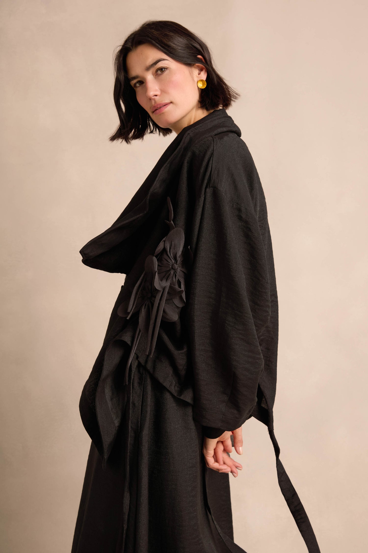 A woman with short dark hair wears the Flower Detail Jacket—a black draped style with bow accents—standing against a beige background.