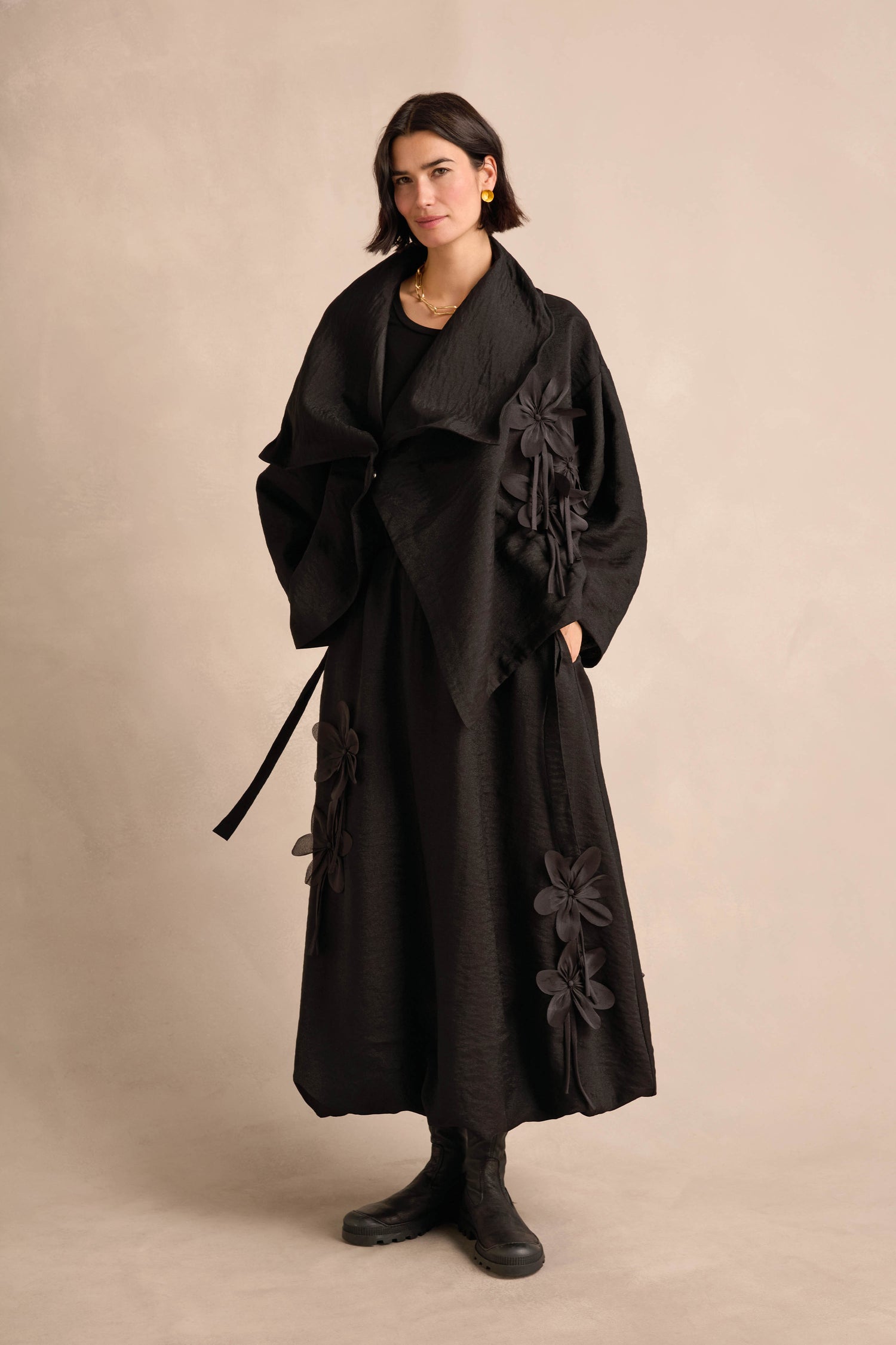 A woman wearing the Flower Detail Jacket in black, paired with a long dress featuring large fabric flower accents, stands against a neutral background.
