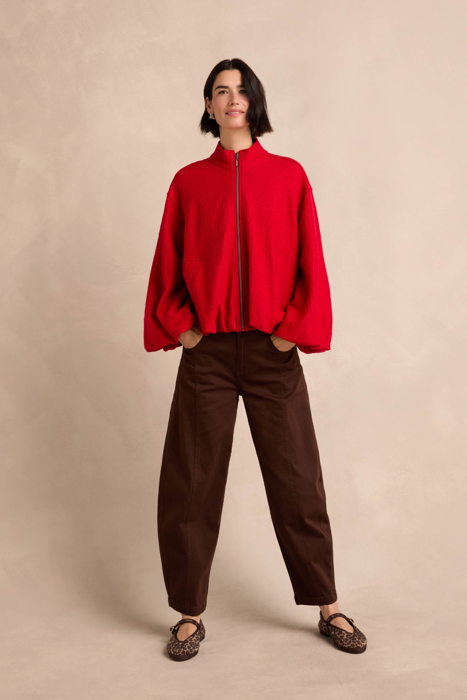 A woman stands with her hands in her pockets, wearing the Puffball Boiled Wool Jacket in red, paired with brown loose pants and leopard-print flats against a neutral background.